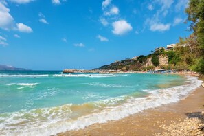 Ein Küstenstrand mit klarem türkisblauem Wasser, einem Kiesstrand und Gebäuden an einem Hügelhang.