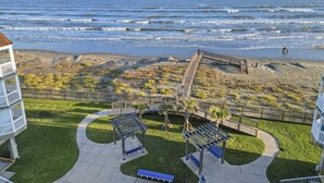 Vista sulla spiaggia dal balcone di un hotel, con una passerella in legno, zone verdi e cielo sereno.
