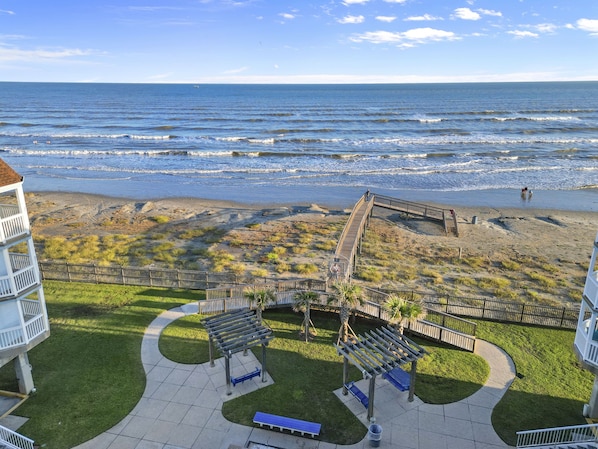A beach view from a hotel balcony with a wooden boardwalk, grassy areas, and a clear sky.