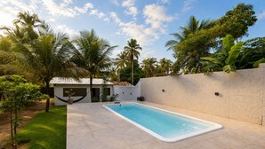 Uma piscina retangular com um deck ladrilhado, rodeada por um muro baixo e vegetação exuberante, incluindo palmeiras.
