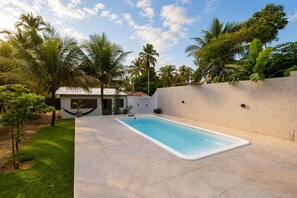 A rectangular swimming pool with a tiled deck, surrounded by a low wall and lush greenery including palm trees. - Casa com Piscina Pertinho da Praia do Boqueir o (Japaratinga)