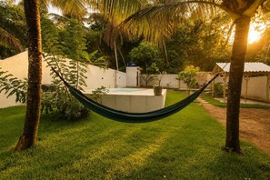 A hammock in a grassy area with a concrete structure, palm trees, and a white building in the background. - Casa com Piscina Pertinho da Praia do Boqueir o (Japaratinga)