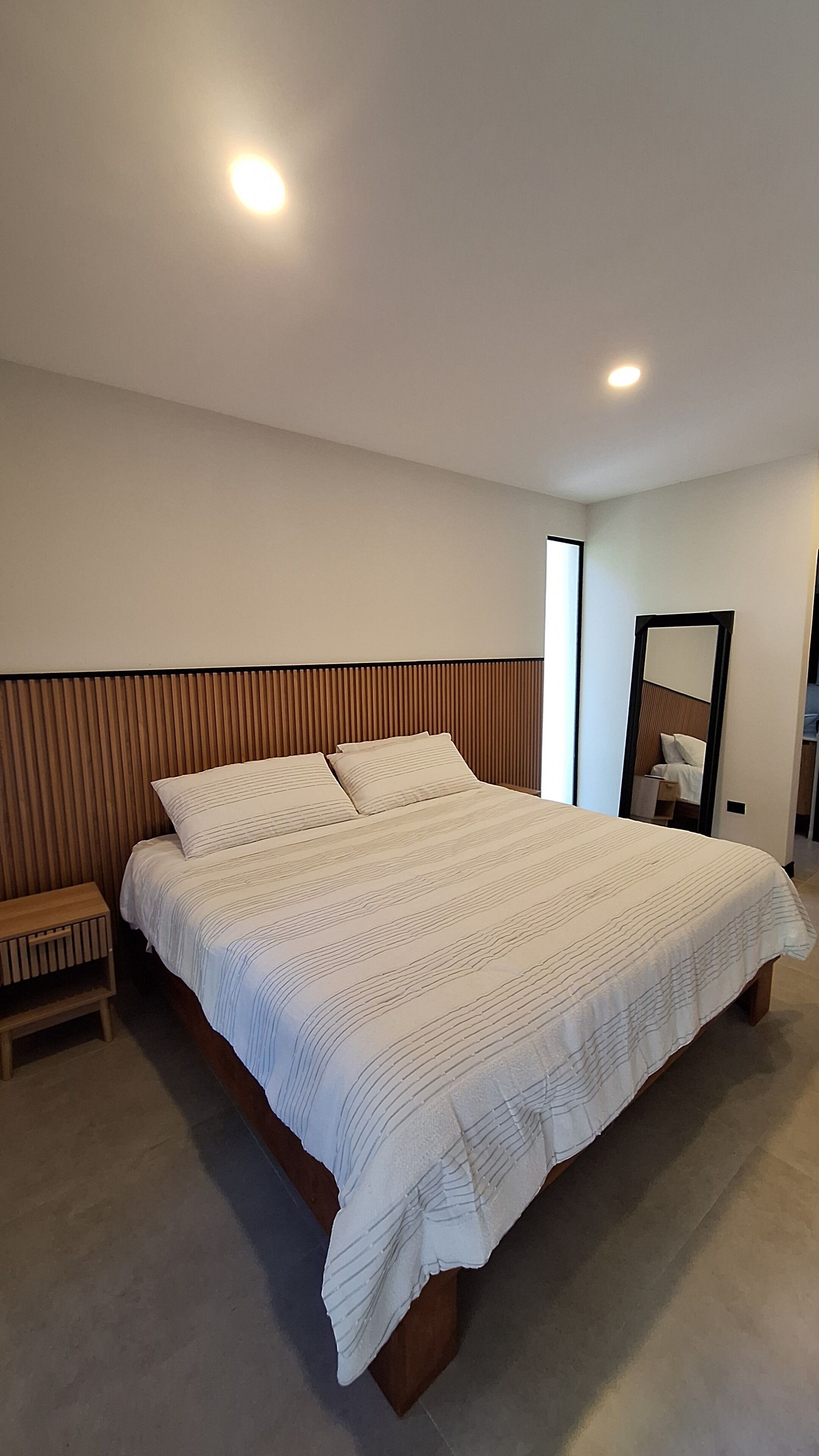 A modern bedroom with a large bed, a nightstand, a mirror, and recessed ceiling lights.