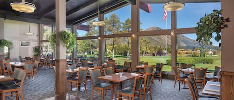 A spacious restaurant with wooden tables and chairs, large windows offering a view of a golf course and mountains.