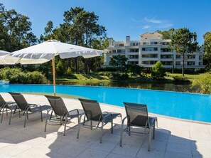 Zona junto a la piscina con sillones de descanso y sombrillas, una piscina azul cristalina y un edificio con varios balcones al fondo.