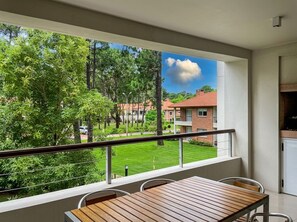 Un balcón con una mesa y sillas de madera, vista a un césped verde y un edificio de ladrillo al fondo.