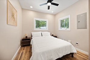 A bedroom with a bed, two bedside tables, a ceiling fan, and two windows with a view of trees.