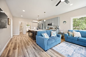 A modern kitchen with grey cabinets, a microwave, and a coffee machine. There are two blue sofas with patterned cushions, a wooden floor, and a large window with a view of trees.