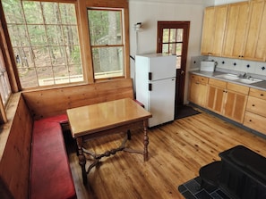 Une cabane avec un banc en bois, une table, un réfrigérateur, des placards en bois et une fenêtre donnant sur les arbres.