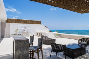 Eine Dachterrasse mit Sitzgelegenheiten im Freien, einer Steinsäule und Blick aufs Meer.
