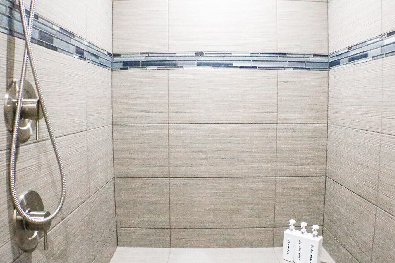 A shower area with tiled walls, a handheld showerhead, and a bottle of shampoo.