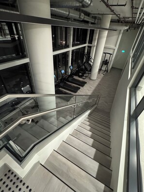 A modern indoor staircase with glass railings, a fitness area with treadmills, and a view of the city at night. - Waterfront luxurious 2-bedroom/2 bath apartment in fabulous Kelowna (Kelowna)