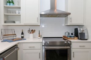 Una cocina con armarios blancos, una campana extractora de acero inoxidable, una cafetera y una botella de vino.