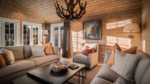 Ein gemütliches Blockhaus-Interieur mit Holzbalkendecke, Kamin, einer Couch, einem Sessel und einem Esstisch.