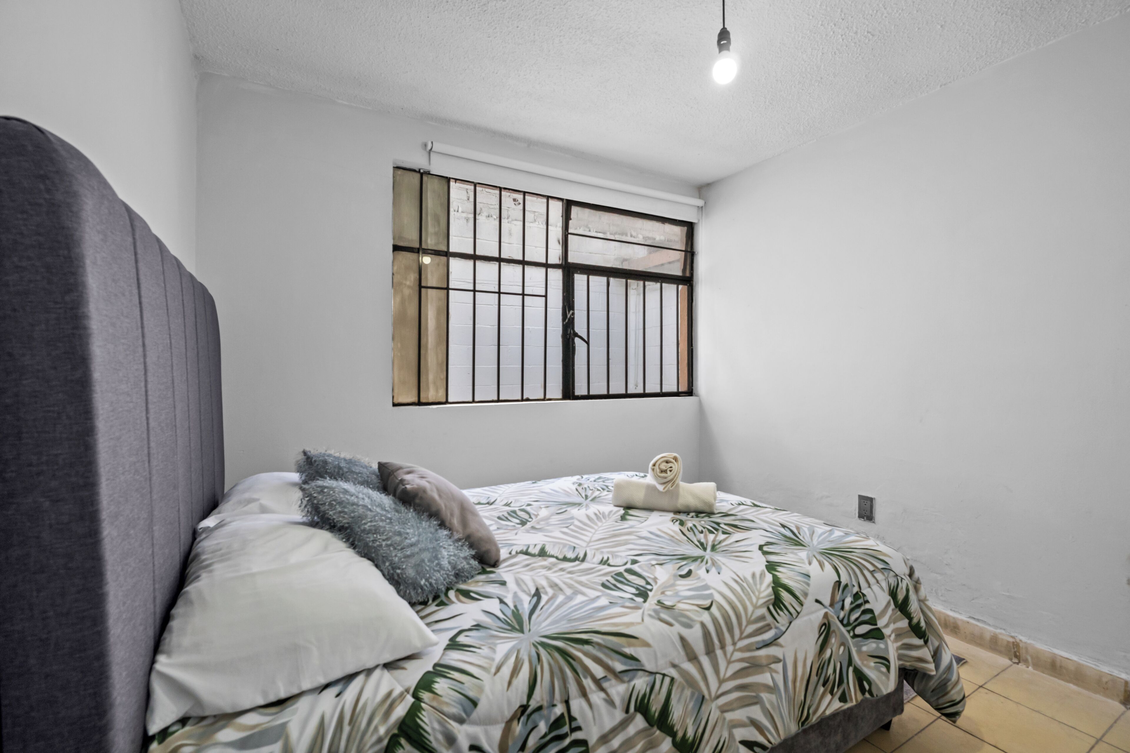 Een slaapkamer met een bed, kussens, een raam met tralies en een lamp.