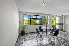 Una habitación con suelo de baldosas, una mesa redonda con sillas, una planta en maceta y una ventana con vistas a zonas verdes y un edificio.