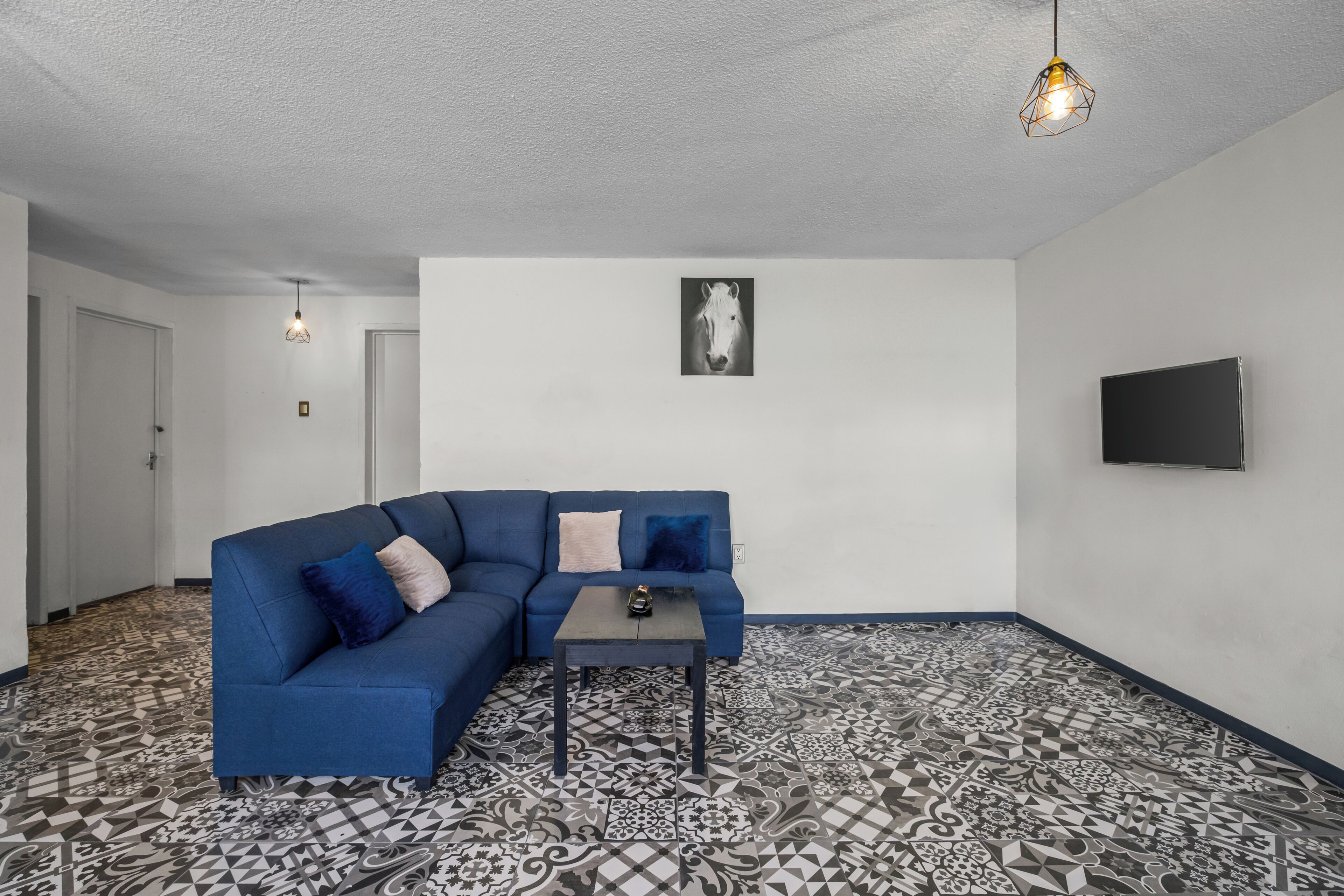 Una habitación con un sofá seccional azul, una mesita, baldosas de suelo estampadas, un televisor montado y una lámpara en el techo.