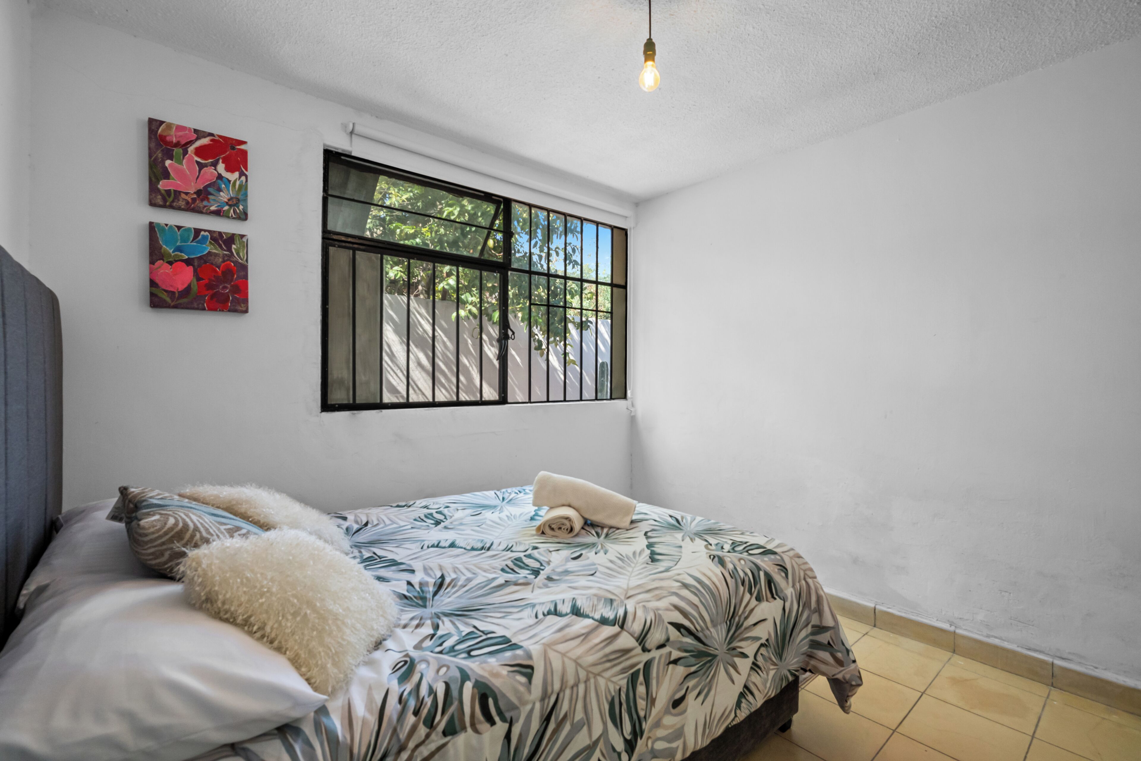 A bedroom with a bed, pillows, a window with bars, and two wall-mounted paintings.