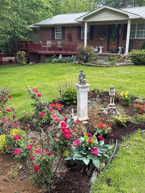 A residential garden with a brick house, a wooden deck, and various flowering plants. - Perfect 2-bedroom house w/ AC in peaceful Tracy City on The Mountain Goat Trail (Tracy City)
