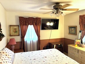 Ein Schlafzimmer mit Bett, Deckenventilator, Fernseher, Kommode und einem Fenster mit Vorhängen.