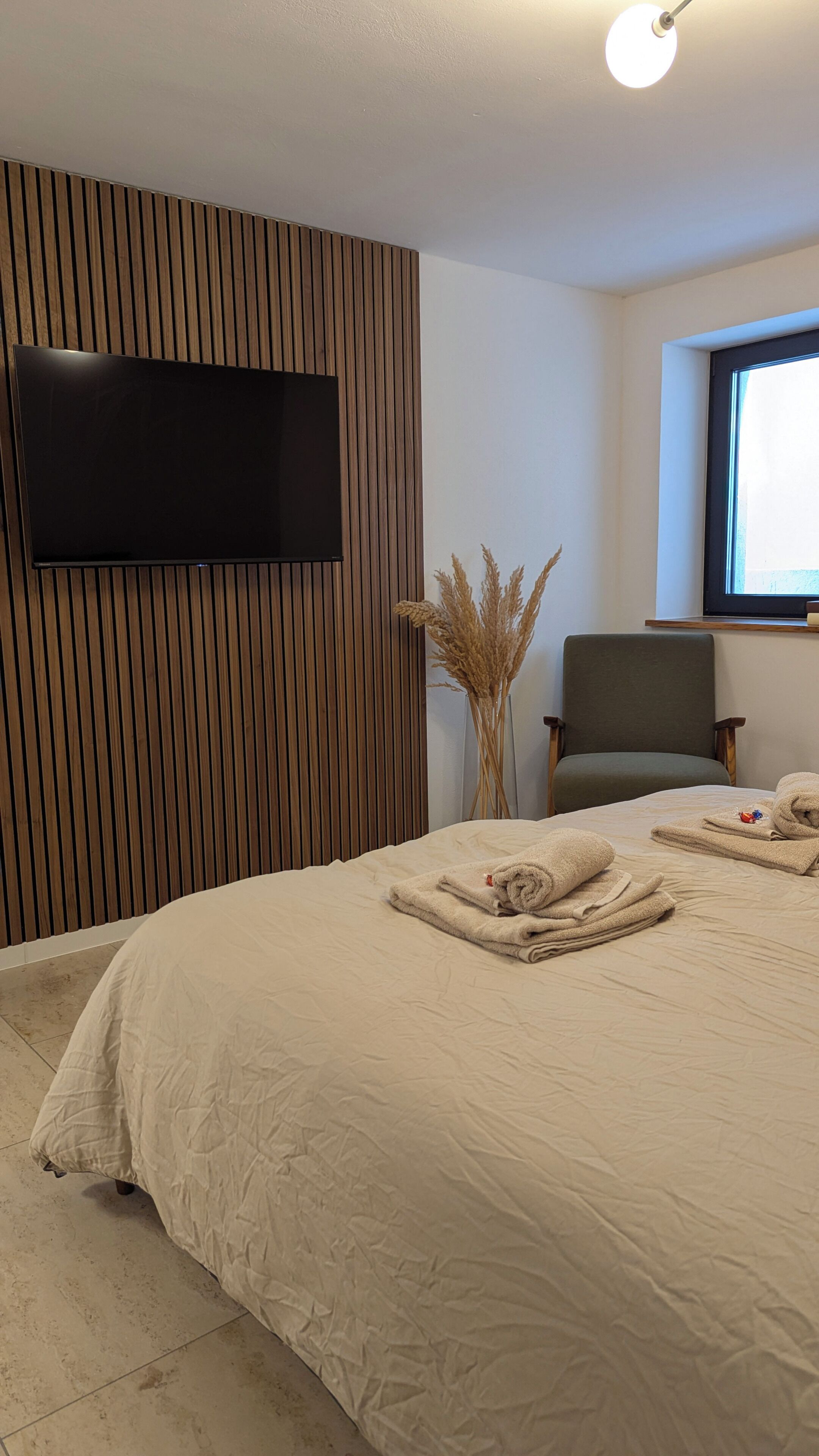 A hotel room with a large bed, a flat-screen TV mounted on a wooden panel wall, a chair, and a window.