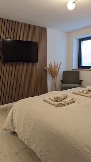 A hotel room with a large bed, a flat-screen TV mounted on a wooden panel wall, a chair, and a window. - MarMys Retreat - Unwind in Nature with Style and Charm (Derental)