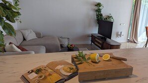 Uma sala de estar moderna com um sofá, uma mesa de centro, uma televisão e um espaço de refeições com um livro, uma tábua de corte e frutas cítricas.