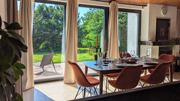 Uma sala de jantar com uma mesa posta para quatro pessoas, vista para o jardim através de grandes janelas e um relógio na parede.