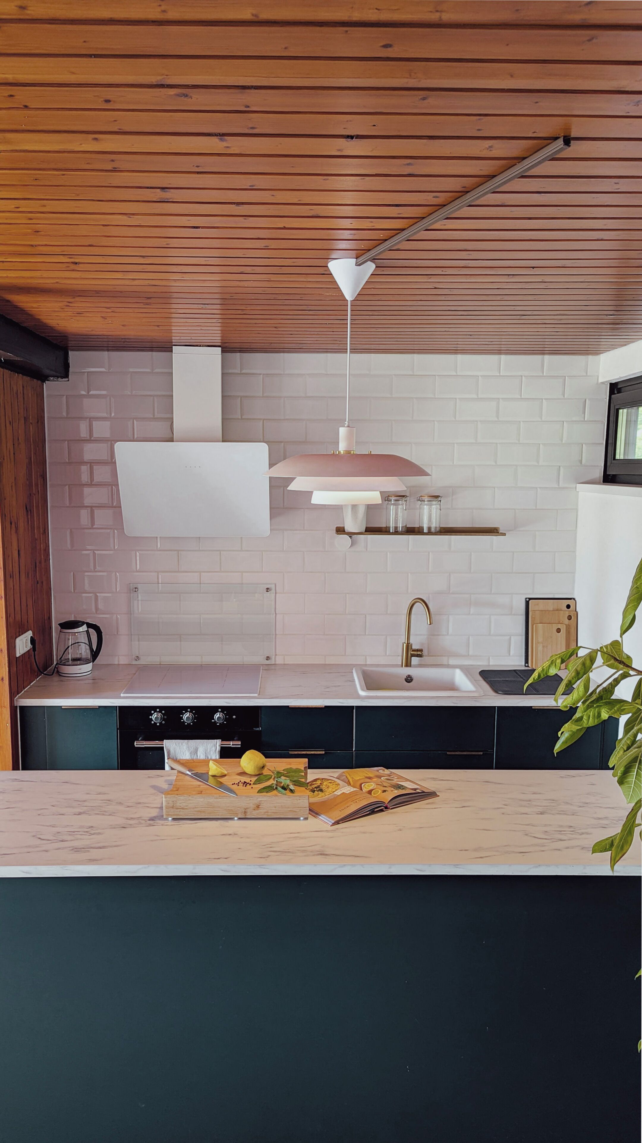 A kitchen with a wooden ceiling, white brick backsplash, a sink, a stove, and a countertop with a cutting board and a book.