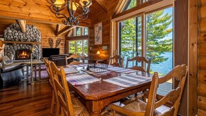 A wooden interior with a fireplace, dining table, chairs, and large windows overlooking a body of water. - Luxury log cabin on Spider Lake in Manitowish Waters (Manitowish Waters)