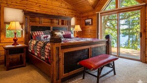 A wooden cabin bedroom with a bed, bench, two nightstands, and a large window. - Luxury log cabin on Spider Lake in Manitowish Waters (Manitowish Waters)