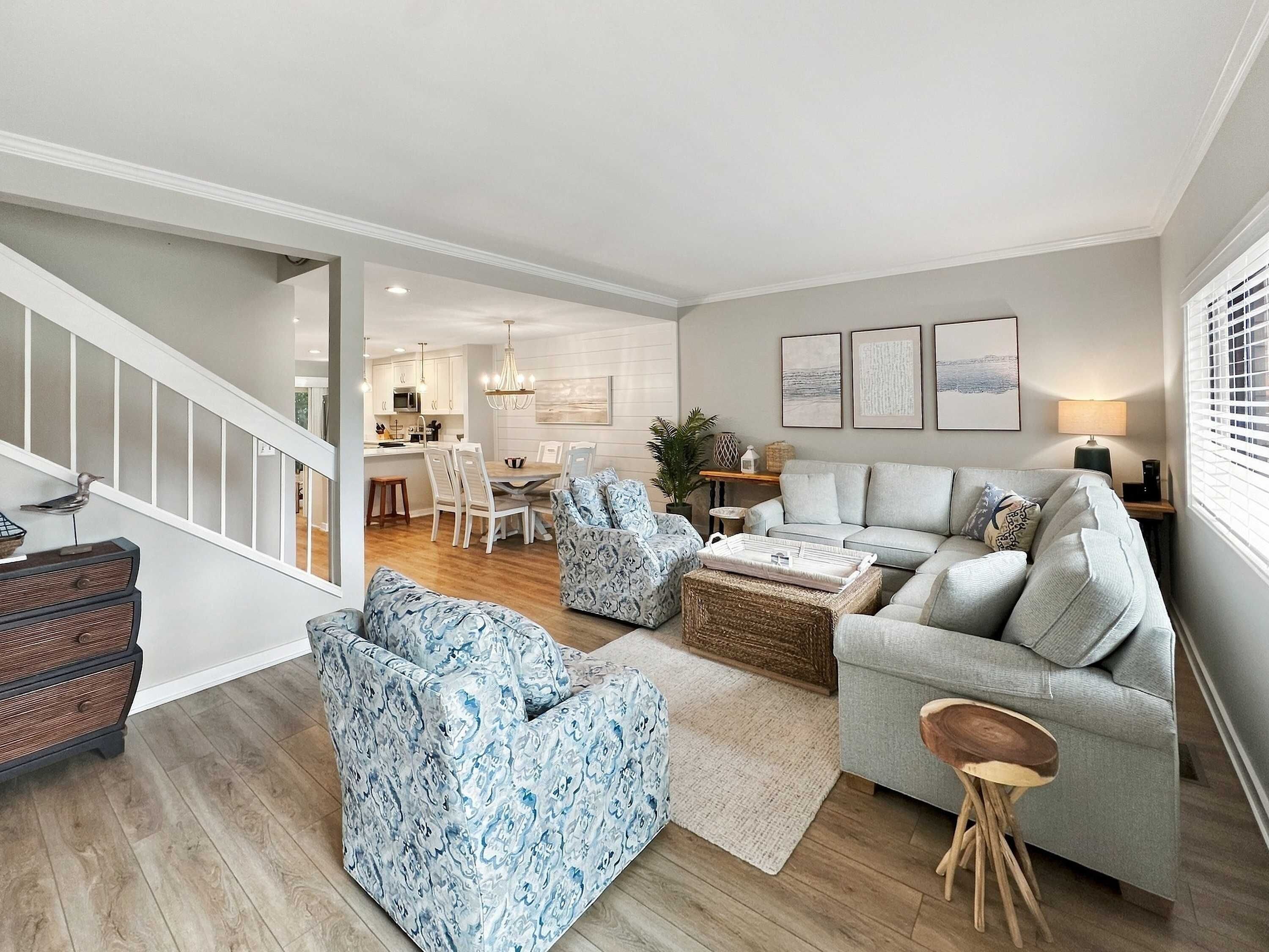 Ein modernes Wohnzimmer mit einem grauen Sofa, gemusterten Sesseln, einem Couchtisch und einer Treppe, die in einen Essbereich führt.