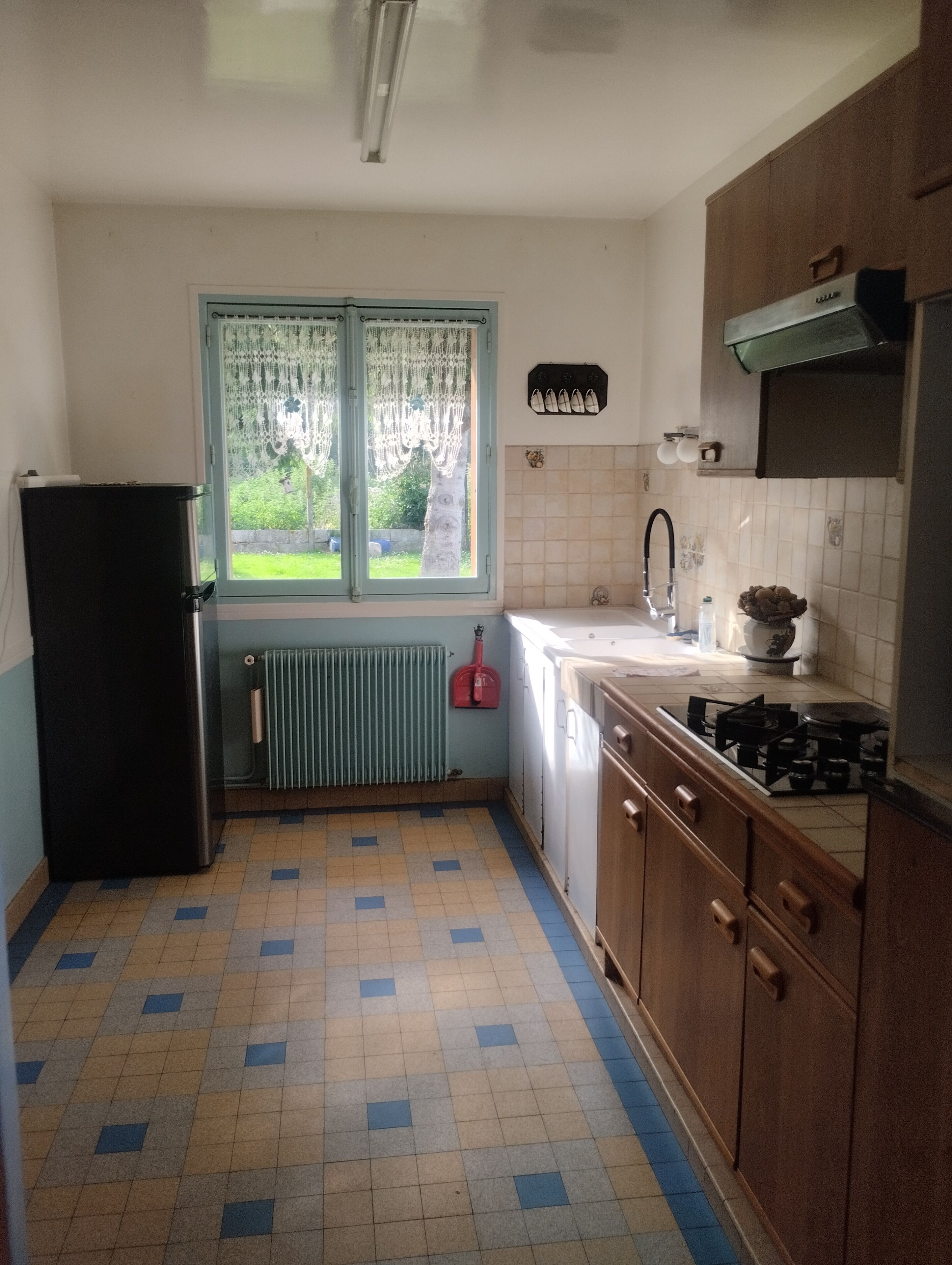 Een keuken met raam, radiator, spoelbak, fornuis en keukenkastjes.