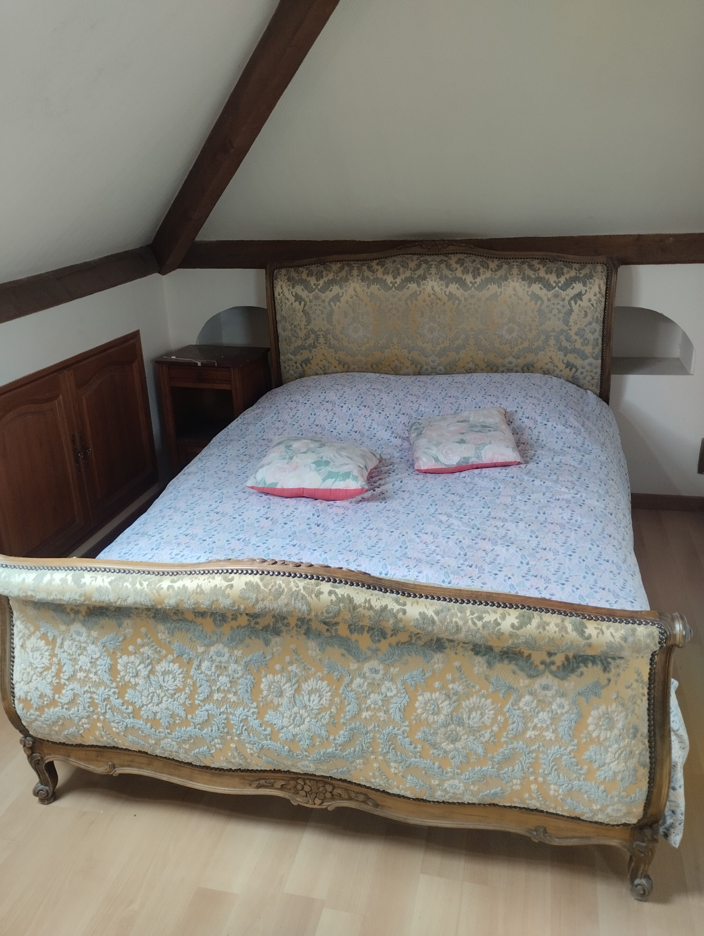 Un lit avec une couvre-lit à motifs floraux et deux oreillers, une table de chevet en bois et une armoire en bois.
