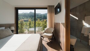 Une chambre d’hôtel moderne dotée d’un grand lit, d’un fauteuil, d’une télévision et offrant une vue sur un paysage boisé.