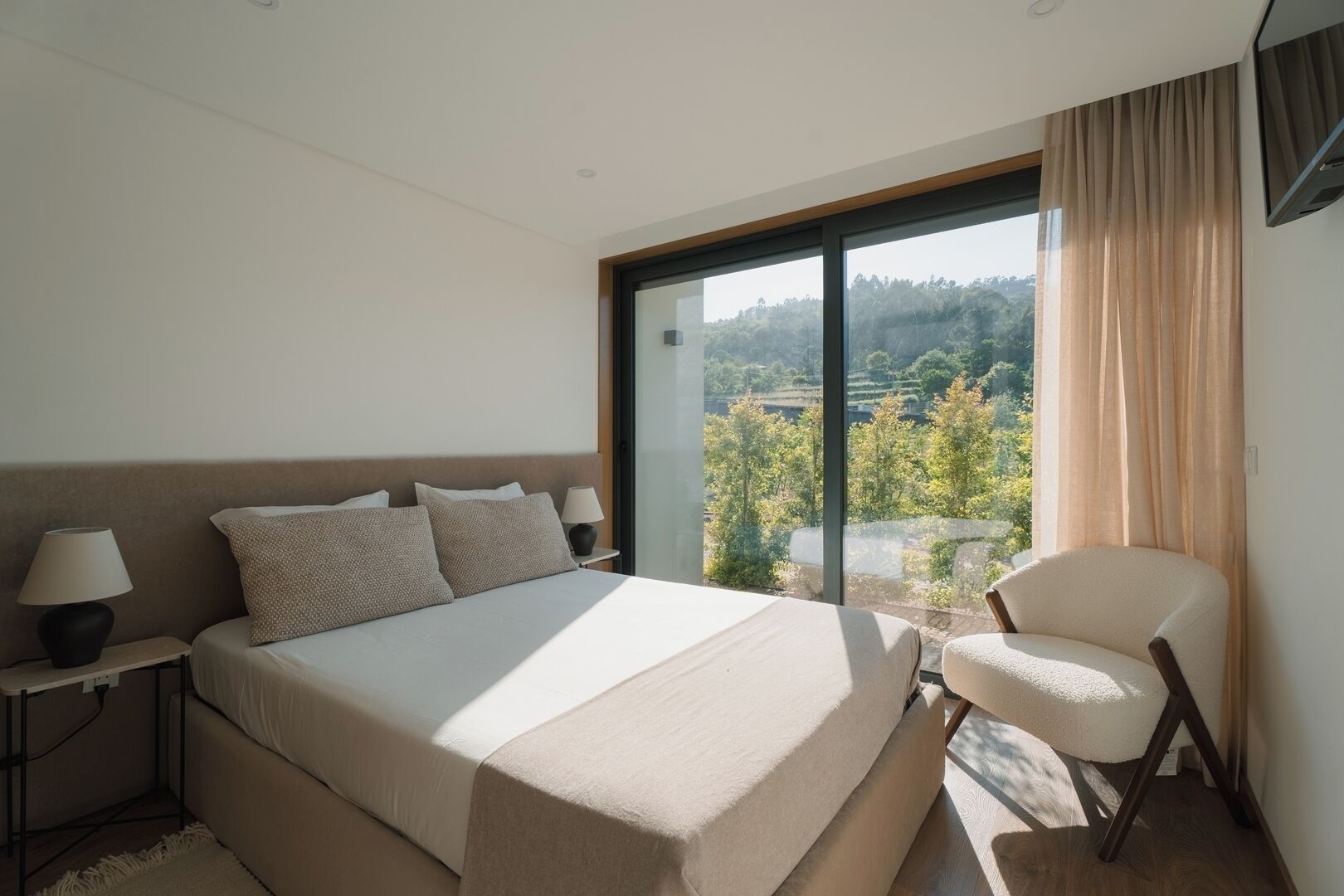 Ein Schlafzimmer mit einem großen Bett, einem Sessel, einem Nachttisch und Blick auf Grün durch ein Fenster.