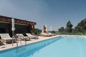 A pool area with lounge chairs and umbrellas, a covered seating area, and a view of trees and hills.
