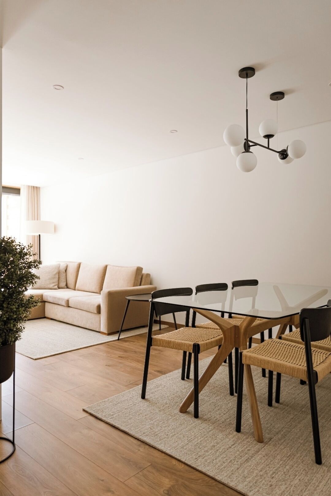 Ein modernes Wohnzimmer mit einem Glastisch, schwarzen Stühlen, einem beigen Sofa, einer Topfpflanze und einer Pendelleuchte.