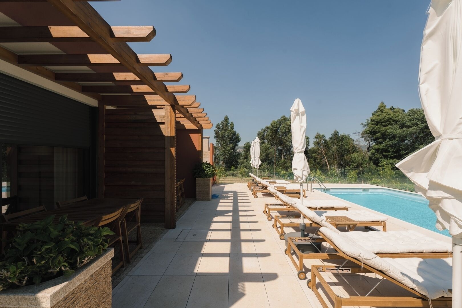 A poolside area with wooden loungers, white umbrellas, and a pergola structure.