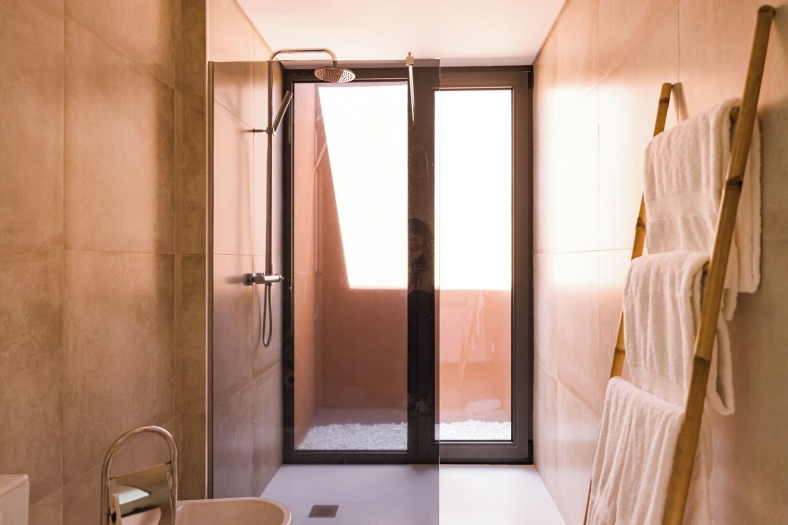 A modern bathroom with a glass shower enclosure, a wooden ladder with towels, and a view through the glass door.