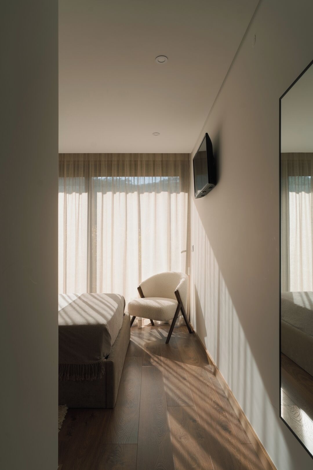 A bedroom with a bed, a chair, a TV mounted on the wall, and a large mirror.