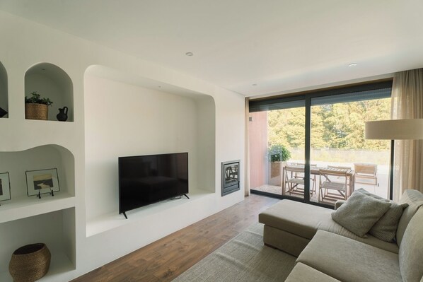 A modern living room with a flat-screen TV, a sofa, a dining area with a table and chairs, and a large sliding glass door leading to an outdoor patio.