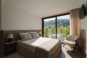 Ein modernes Schlafzimmer mit einem großen Bett, einem Sessel, einem Fernseher und Blick auf eine Landschaft.