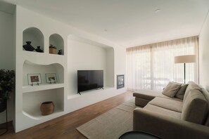 Ein modernes Wohnzimmer mit Flachbildfernseher, einer Couch, einem Couchtisch und einem großen Fenster mit durchsichtigen Vorhängen.