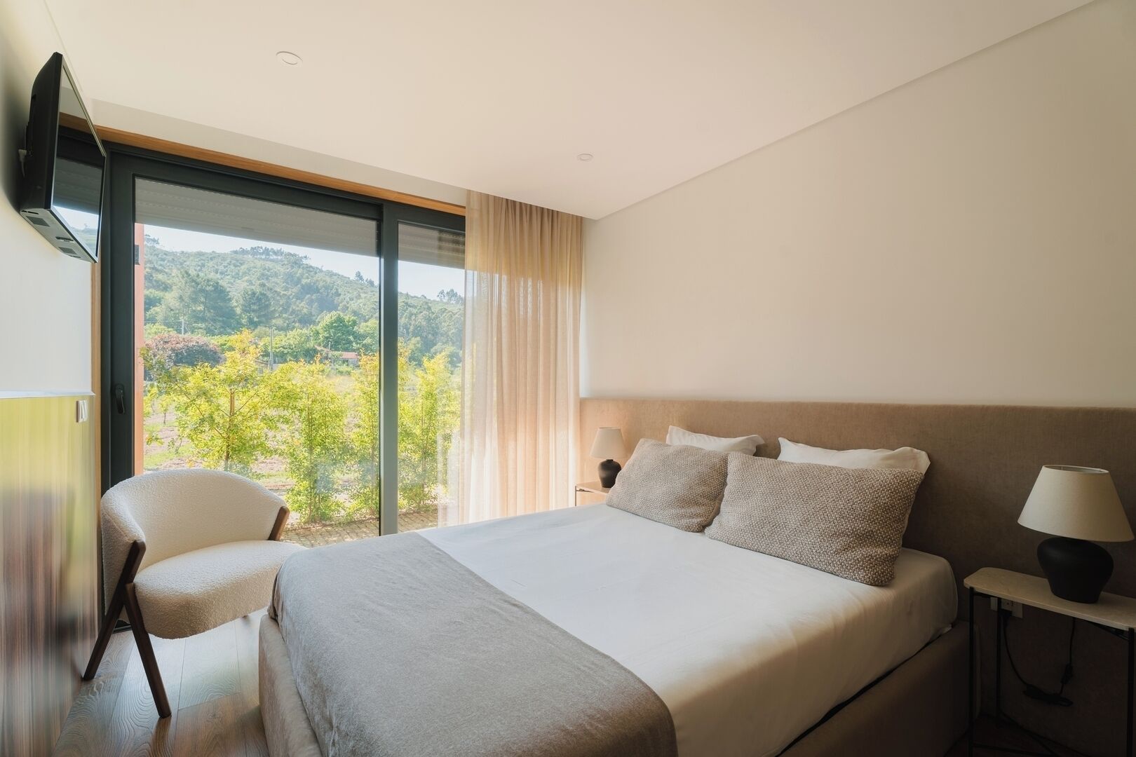 Hotelkamer met een groot bed, een stoel, een nachtkastje, een lamp en uitzicht op groen door een raam.