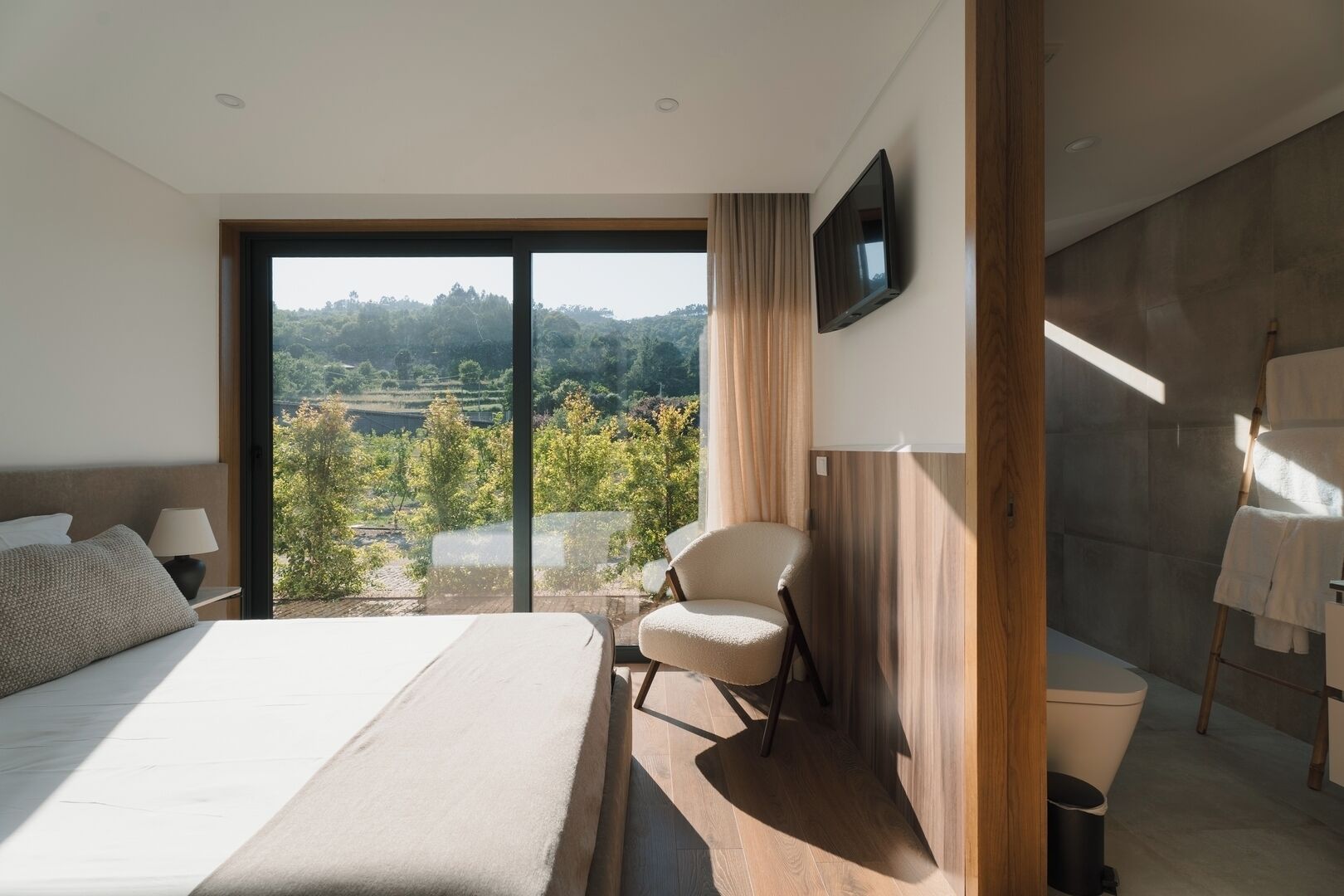 Habitación de hotel moderna con una cama grande, una silla, un televisor y vista a un paisaje boscoso.