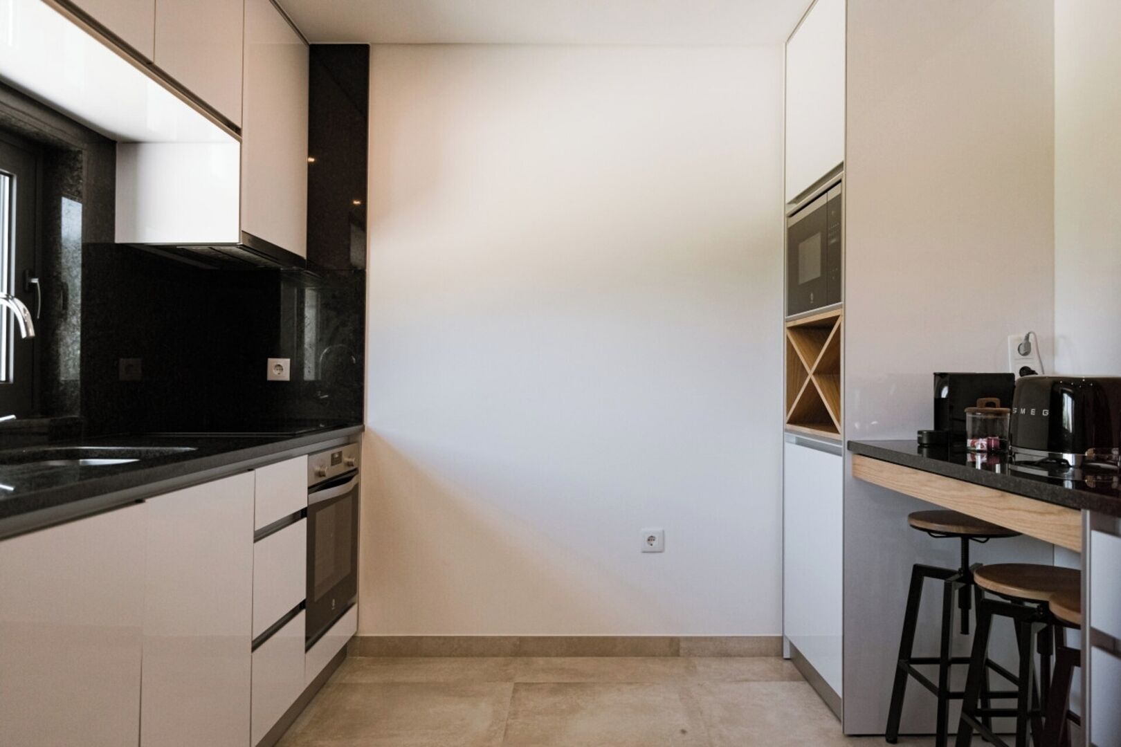 Uma cozinha moderna com bancadas pretas, armários brancos e forno embutido. Possui um espaço de bar com banquetas e uma máquina de café.