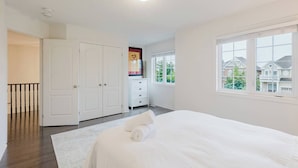 A bedroom with a large bed, a wardrobe, a dresser, and a window with a view of houses.