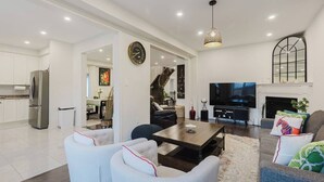A modern living room with a sofa, coffee table, and TV. There is a kitchen with a refrigerator and cabinets. - Spacious 4BR Retreat | Backyard | Near Ontario (Oshawa)