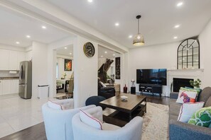 A modern living room with a sofa, coffee table, and TV. There is a kitchen with a refrigerator and cabinets. - Spacious 4BR Retreat | Backyard | Near Ontario (Oshawa)